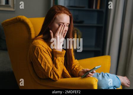 Das Porträt einer müden jungen Frau mit verschwommenem Sehen besteht darin, beim Tippen mit dem Smartphone die Augen zu reiben und zu blinzeln. Stockfoto