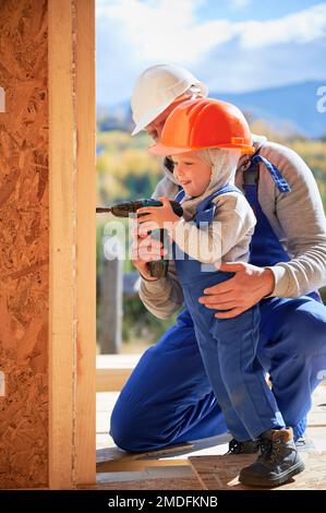 Vater mit Kleinkind Sohn Gebäude Holzrahmen Haus. Baumeister und Kind mit Schraubendreher auf der Baustelle, an sonnigen Tagen mit Helm und blauen Overalls. Zimmerei- und Familienkonzept. Stockfoto