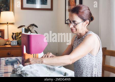 Eine Frau mittleren Alters, die zu Hause in ihrem Wohnzimmer eine Nähmaschine benutzt, um ein Kleid zu machen Stockfoto