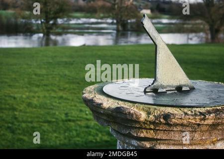 Eine alte Sonnenuhr aus Metall, die auf einem Steinsockel oder Sockel auf einem grasbedeckten Rasen mit einem See im Hintergrund aufgestellt ist Stockfoto