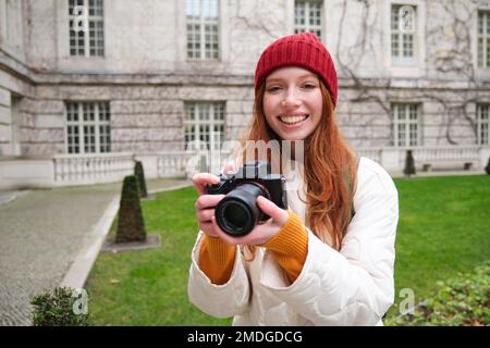 Eine rothaarige Fotografin macht Fotos mit einer professionellen Kamera im Freien, macht Straßenaufnahmen, sieht begeistert aus, während sie Fotos macht Stockfoto