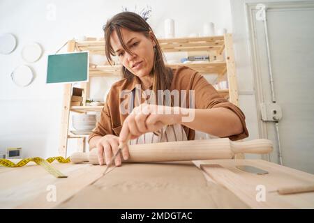 Eine Frau in einer Töpferwerkstatt schneidet Ton, während sie einen Stift hält. Kunsthandwerkstatt Stockfoto