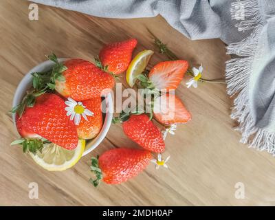 Blick auf ganze und halbierte Erdbeeren in einer weißen Schüssel und auf einem Holztisch, dekoriert mit Zitronenscheiben und Blütenblättern, dekorative Fabrik Stockfoto