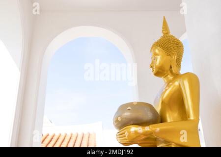 Ein goldener Buddha mit einer Almosenstatue an einem alten Stupa vor einem weißen Bogen und blauem Himmel im Hintergrund. Thailand. Stockfoto