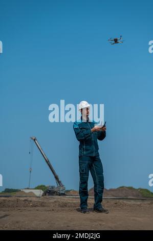 Ein Mann in Helm und Overall steuert eine Drohne auf einer Baustelle. Der Bauherr führt die technische Überwachung durch. Stockfoto