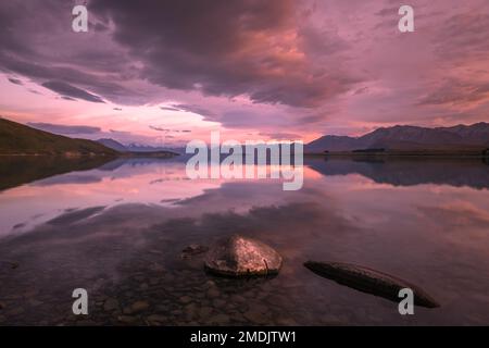 Sonnenaufgang über einem spiegelruhigen Lake Tekapo und schneebedeckten Bergen in Neuseeland mit Felsen im Vordergrund Stockfoto