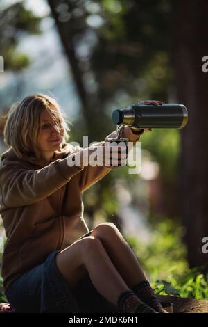 Eine Frau, die in einem Park sitzt, gießt gerne heißes Wasser aus einer Thermoskanne in einen Mate Becher. Stockfoto