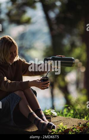 Eine Frau, die in einem sonnigen Park sitzt, gießt heißes Wasser aus einer Thermoskanne in einen Mate Becher. Stockfoto