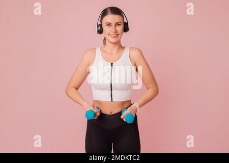 Fröhlich lächelnde junge Frau mit blauen Kurzhanteln über pastellrosa Hintergrund. Studioporträt einer süßen, fröhlichen Fitnessfrau, die Sportbekleidung beim Sport trägt Stockfoto