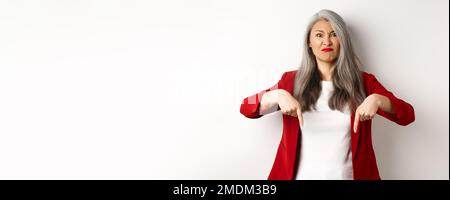 Enttäuschter und wütender asiatischer Arbeitgeber, der die Finger nach unten zeigt, unzufrieden und aufgeregt aussieht und über weißem Hintergrund steht Stockfoto