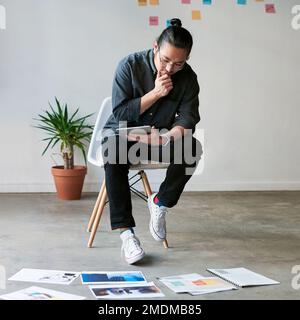 Alle Aspekte ernsthaft zu berücksichtigen. Gesamtbild eines kreativen jungen Geschäftsmanns, der auf einem Stuhl sitzt und sein digitales Tablet bei verwendet Stockfoto