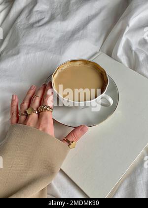 Eine vertikale Aufnahme einer Hand, die eine Tasse Kaffee auf einem Buch berührt Stockfoto