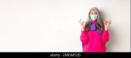 Covid-19, Quarantäne- und Sperrkonzept. Überrascht und beeindruckt asiatische ältere Frau in medizinische Maske suchen fasziniert, Lob etwas Gutes und Stockfoto