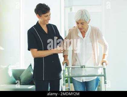 Bitte helfen Sie mir. Eine attraktive junge Gesundheitsfachkraft, die ihrem Seniorpatienten hilft, mit einer Gehhilfe in einem Pflegeheim zu gehen. Stockfoto