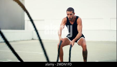 Das ist eine Schlacht, die er unbedingt gewinnen will. Ein gutaussehender junger Sportler, der tagsüber im Freien mit Kampfseilen trainiert. Stockfoto