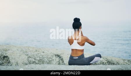 Es ist machtvoll, still zu bleiben. Rückblick auf eine nicht erkennbare Frau, die quer über den Beinen sitzt und dabei allein am Meer meditiert Stockfoto