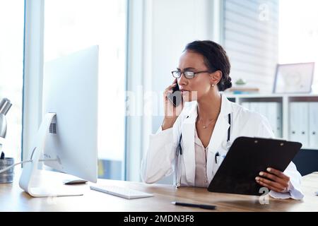 Ich würde Sie gerne für eine Nachuntersuchung sehen. Eine junge Ärztin, die ein Smartphone und einen Computer in ihrem Sprechzimmer benutzt. Stockfoto