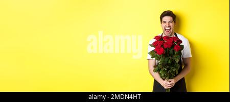 Lustiger junger Verkäufer in schwarzer Schürze, der Strauß Rosen hält, Blumenhändler lachend und über gelbem Hintergrund stehend Stockfoto