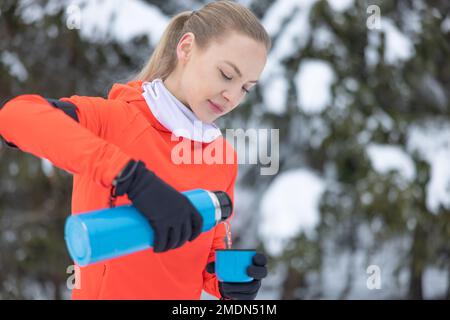 Eine junge Frau in warmer Fitnesskleidung gießt heißen Tee in eine Tasse aus Metallthermoskannen, nachdem sie im Winter auf dem Sportplatz trainiert hat. Stockfoto
