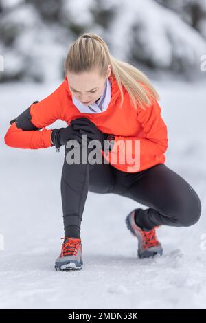 Eine junge, fit geschnittene Frau hält ein verstärktes Knie, nachdem sie ihr Kreuzband während des Langlauftrainings im Schnee während der Wintersaison gestrafft hat. Stockfoto