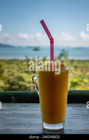 Frisches tropisches Mangogetränk mit einem Strohhalm auf einem Restauranttisch mit Aussicht. In Ko Lanta, Krabi, Thailand Stockfoto