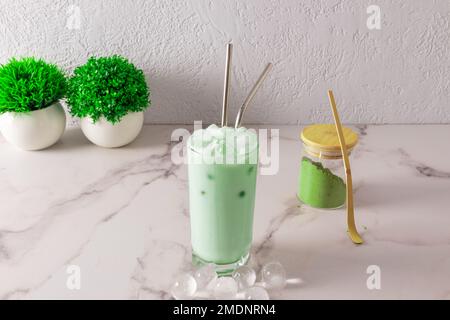 grüner matcha-Dalgon-Tee in einem großen Glas mit Metallstroh und runden Eisstücken. Grüner Tee-Pulver in einem Glasgefäß. Eine Alternative zu kaltem Kaffee Stockfoto