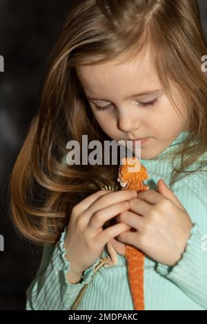 Porträt eines hübschen Mädchens mit rotem Agama-Leguan auf grauem Hintergrund. Kleines Kind, das mit Reptilien spielt. Selektiver Fokus. Hochwertiges Foto Stockfoto