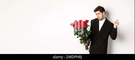 Bild von eleganten und frechen Mann in schwarzem Anzug, selbstbewusst und hält Strauß von roten Rosen, gehen auf ein romantisches Date, stehen gegen weiß Stockfoto