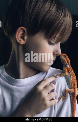 Porträt eines Jungen mit rotem Agama-Leguan auf grauem Hintergrund. Kleines Kind, das mit Reptilien spielt. Selektiver Fokus. Hochwertiges Foto Stockfoto