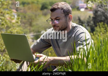 Junger erwachsener Mann mit Brille, der auf grünem Gras sitzt und an einem Laptop arbeitet. Stockfoto