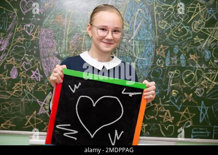 Eine Brillenschülerin im mittleren Schulalter hält ein Schieferbrett, auf das ein Herz gezeichnet wird. Stockfoto