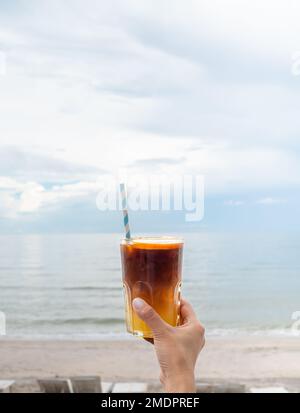 Ein Glas frisch gefrorenes orangefarbenes Ameriano mit handgehaltener ​​orange-Scheibe am Strand, Meereslandschaft, vertikaler Stil. Kalter schwarzer Kaffee mit Stockfoto