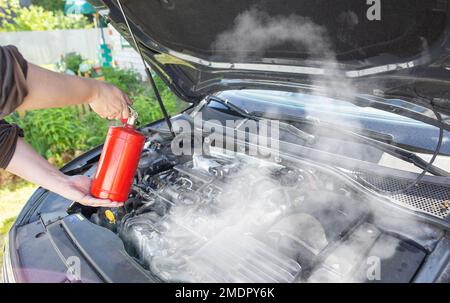 Der Fahrer des Wagens leitet einen Pulverfeuerlöscher an den Motorraum des Wagens, in dem sich Feuer und Rauch befinden. Autofeuer, Hintergrund Stockfoto