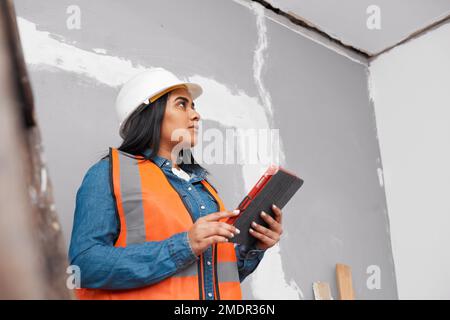 Eine junge, südasiatische Bauarbeiterin verwendet ein Tablet, um ein Projekt zu protokollieren Stockfoto
