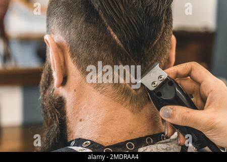 Hipster-Mann im Friseursalon, der Bart und Haare schneidet - Friseurin, die Haarschneider verwendet, um mit geometrischer Zeichnung auf dem Nacken zu schneiden - B Stockfoto
