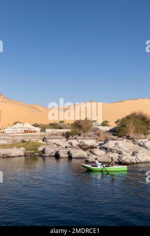 ASSUAN, ÄGYPTEN - 29. Dez. 2022. Vertikale wunderschöne Landschaft mit zwei nubischen arabern, die am Nilufer rudern, mit Sanddünen-Bergen im Hügel Stockfoto