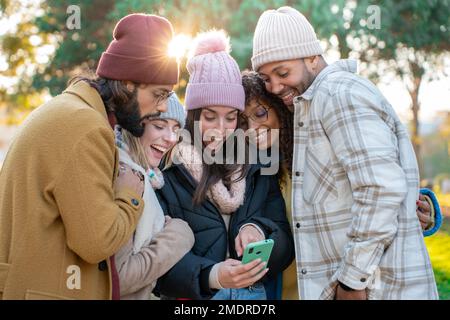 Eine große Gruppe von jungen, multiethnischen Freunden, die auf ihr Handy schauen, sind aufgeregt und haben draußen ein Lächeln auf den Lippen. Stockfoto