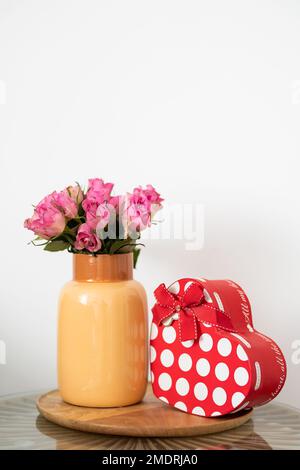 Valentinstagsgeschenk, eine rote herzförmige Box mit weißen Punkten, wird auf einem Holztablett neben einer Vase mit rosa Rosen platziert. Stockfoto