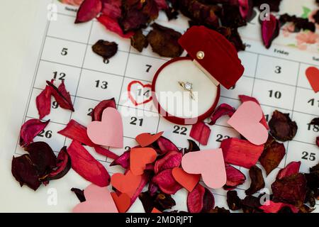 Die Ringbox wird zusammen mit vielen Blütenblättern auf den Kalender gesetzt, und der Valentinstag ist auf dem Kalender rot markiert. Heiratsantrag Stockfoto