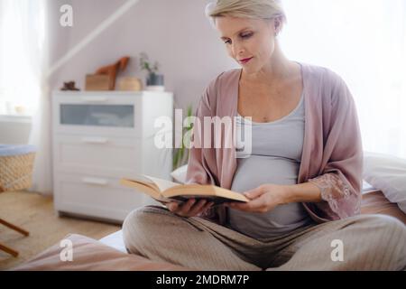 Schwangere Frau, die auf dem Bett sitzt und Buch liest. Stockfoto