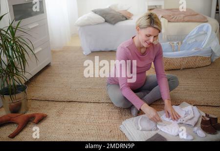 Aus dem Blickwinkel einer schwangeren Frau, die kleine Klamotten für ihr Baby ansieht und sie auf die Entbindungsstation packt. Stockfoto