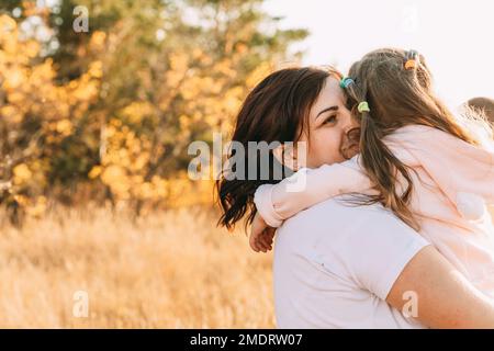 Wunderschöne junge Frau und ihre charmante kleine Tochter umarmen und lächeln draußen Stockfoto