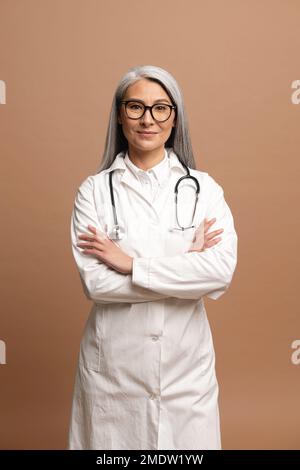 Vertikales Foto der intelligenten professionellen grauhaarigen asiatischen Ärztin, trägt Brille, medizinisches weißes Kleid und Stethoskop, schaut auf die Kamera, Therapeutin Arzt steht isoliert auf braun Stockfoto