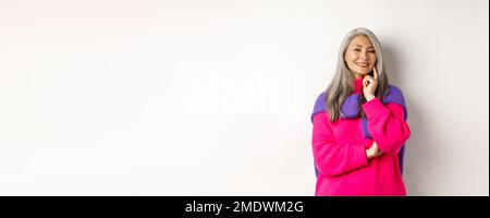 Modische asiatische Frau mittleren Alters mit grauen Haaren, lächelnd zufrieden und nachdenklich, mit einer Idee, stehen über weißem Hintergrund Stockfoto