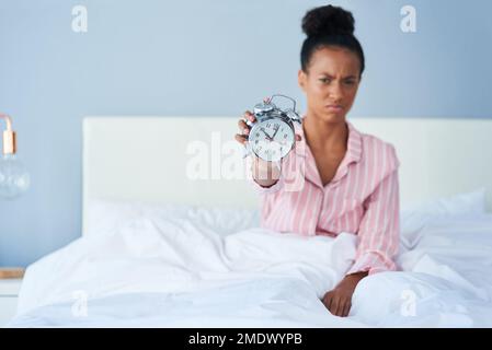 Warum hast du mich so früh geweckt? Eine attraktive junge Frau, die ihren Wecker vor der Kamera im Bett zu Hause hält. Stockfoto