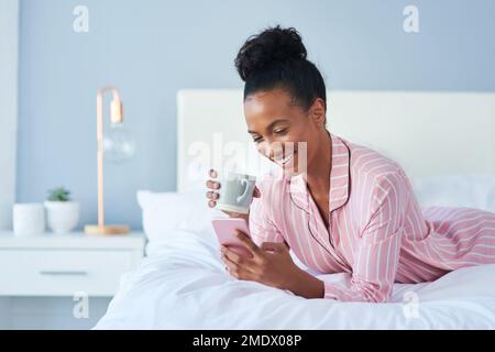 Zeit, etwas heißen Klatsch einzuholen. Eine attraktive junge Frau, die Kaffee trinkt, während sie ihr Handy zu Hause im Bett benutzt. Stockfoto