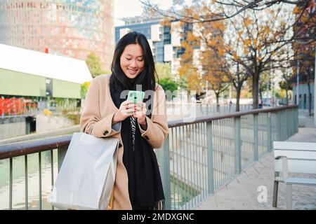 Junge, wunderschöne asiatische Frau lächelt und schreibt eine Nachricht in die Smartphone-App. Porträt eines glücklichen chinesischen Mädchens, das ein Handy benutzt, nachdem er in einer Straße eingekauft hat. Verbindungskonzept. Hochwertiges Foto Stockfoto