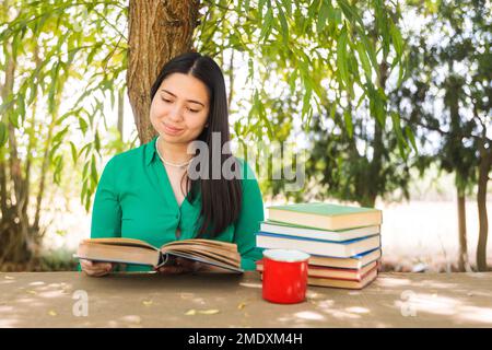 Teenager, die auf dem Feld unter einem Weidenbaum Bücher liest, mit einer Kaffeetasse. Weltbuchtag Stockfoto