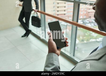 Nahaufnahme einer Hand eines Schwarzen, der ein Handy gegen einen jungen Geschäftsmann in formeller Kleidung hält, der im Büro am Fenster steht Stockfoto