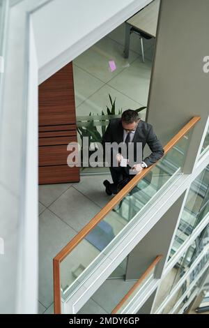 Über dem Bild eines jungen, gut gekleideten Geschäftsmannes, der SMS auf dem Smartphone schickt, während er an transparenten Zaungeländern in einem modernen Bürozentrum steht Stockfoto